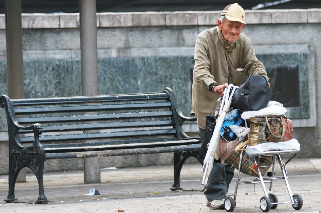 An elderly Japanese man stands near a bench with a trolley