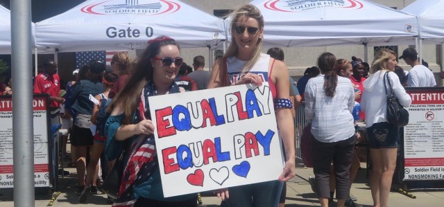 Two soccer fans hold a sign that reads: 'Equal Play Equal Pay'