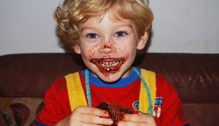 A young boy whose face is covered in chocolate, eating a muffin