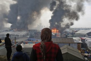 People stand and watch migrant camp Calais on fire.