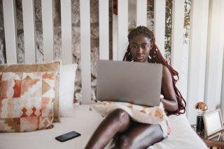 a-portrait-of-a-beautiful-young-black-braided-female-on-the-bed-in-her-apartment-bedroom-working-at_t20_wk6kQ7