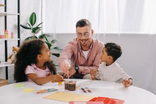 smiling caucasian teacher in eyeglasses and african american kids drawing pictures with paints