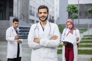 Arabic or hindu man doctor in front of multiethnic team, looking at camera with arms crossed